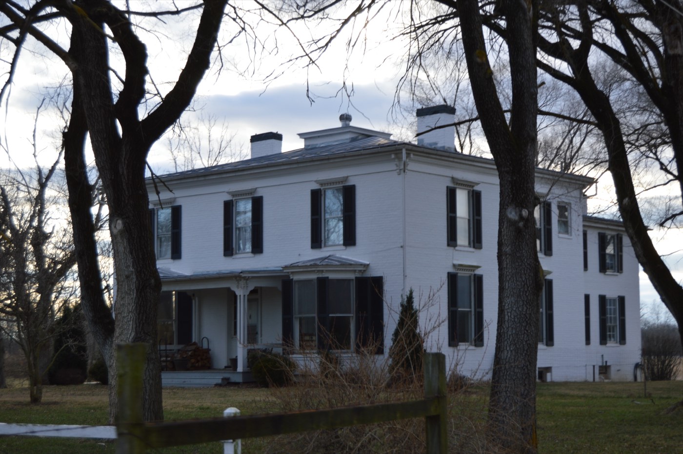 A white two-story farmhouse surrounded by trees and open fields in a rural setting